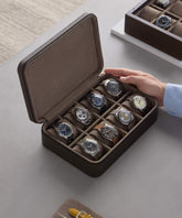 A person unveils the Fraser 8 Watch Case - Brown by TAWBURY, revealing eight exquisite wristwatches. It rests elegantly on a gray surface, with a smaller case subtly peeking from the background—a true treasure for watch collectors.